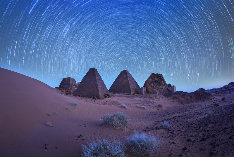Five ancient pyramids in a desert setting with startrails arching in the night sky